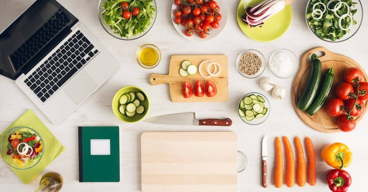 Overhead photo of a kitchen surface covered with a laptop and notebook for meal planning, along with cutting boards, knives, and vegetables.