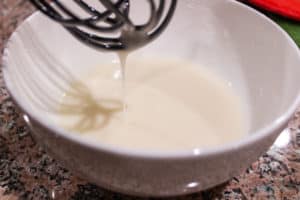 White bowl with lemon glaze in bowl and dripping from whisk above. Bowl is on a kitchen counter.