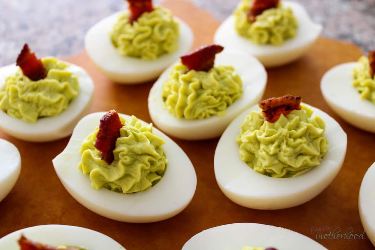 Avocado Deviled Eggs garnished with bacon on a wood serving tray.